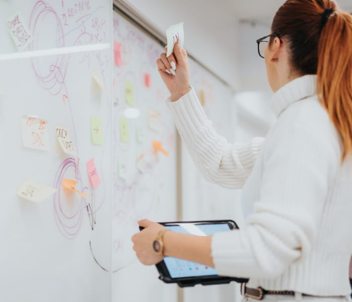 A woman adding a sticky note to the whiteboard while holding a tablet in her hand.
